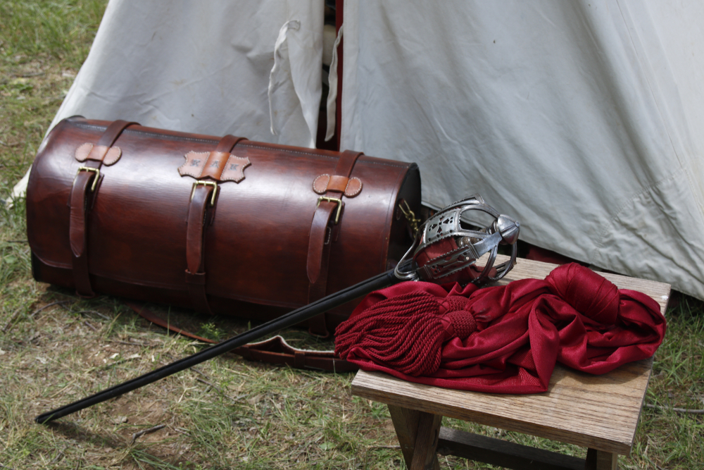 A basket-hilted sword rests on a wooden table with a red sash, beside a leather chest near a canvas tent.