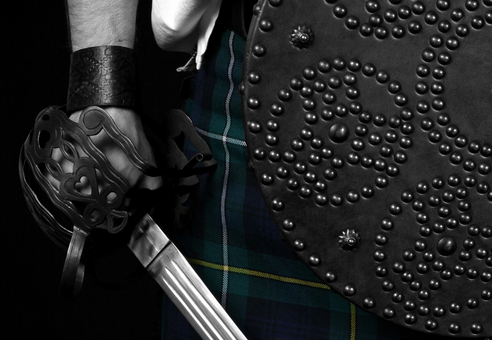 A person in a plaid kilt holds a basket-hilted broadsword beside a studded targe.