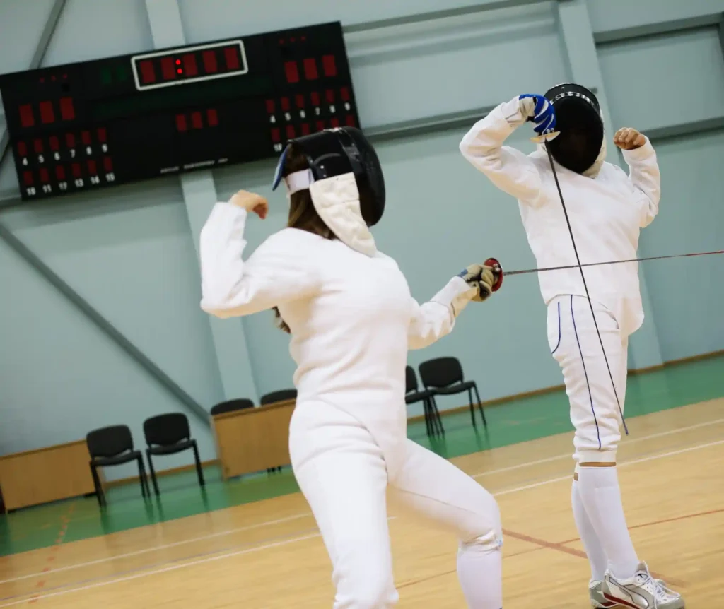 Two fencers in a match, wearing protective gear and engaged in a duel on an indoor court.